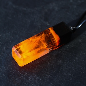 May include: A rectangular pendant featuring a combination of solid black material and translucent orange resin with swirling, flame-like patterns inside. The pendant is attached to a thin black cord, creating a contrast between the dark and bright sections.