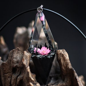 Op de afbeelding: Een heldere, diamantvormige hars hanger met een roze bloem erin. Het onderste gedeelte is zwart en de hanger hangt aan een zwart koord. De hanger wordt getoond tegen een donkere achtergrond en een stuk drijfhout.