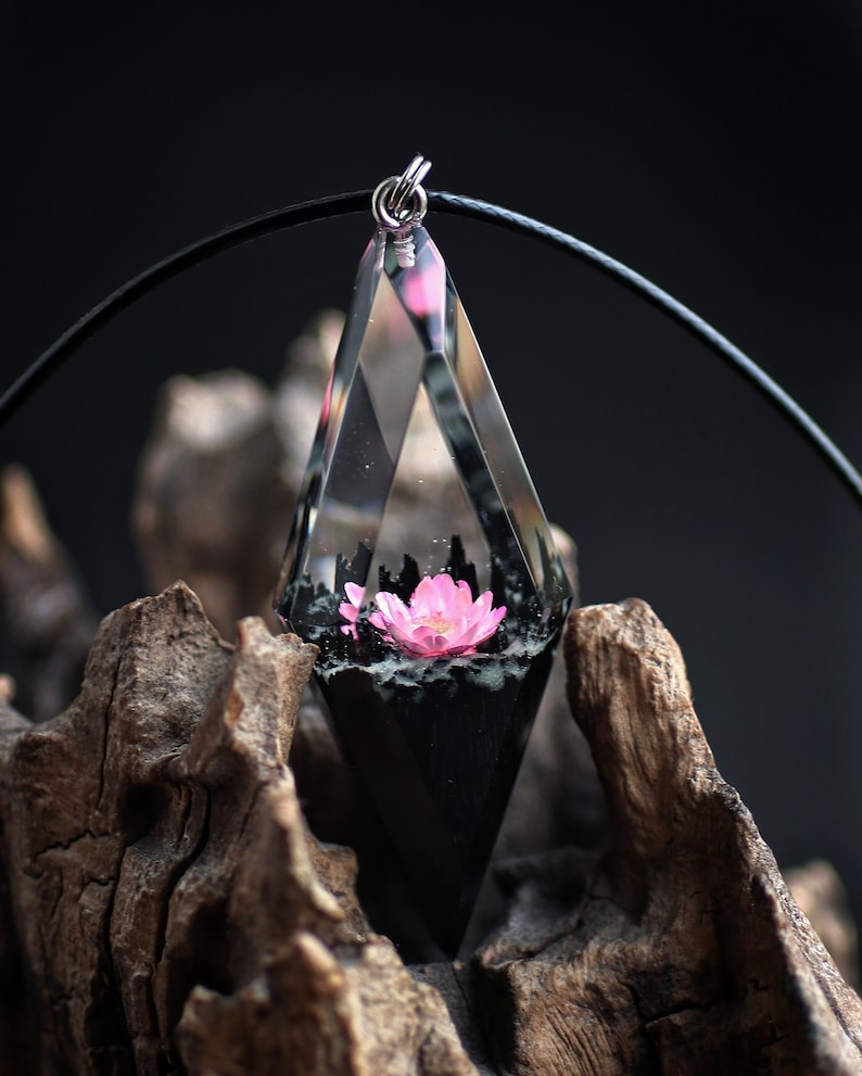 Op de afbeelding: Een heldere, diamantvormige hars hanger met een roze bloem erin. Het onderste gedeelte is zwart en de hanger hangt aan een zwart koord. De hanger wordt getoond tegen een donkere achtergrond en een stuk drijfhout.