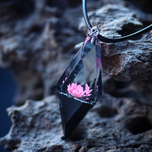 Op de afbeelding: Een geometrische hangerketting met een roze bloem ingekapseld in heldere hars met een zwarte basis. De hanger hangt aan een zwart koord. De ketting wordt getoond op een getextureerd, grijs oppervlak.