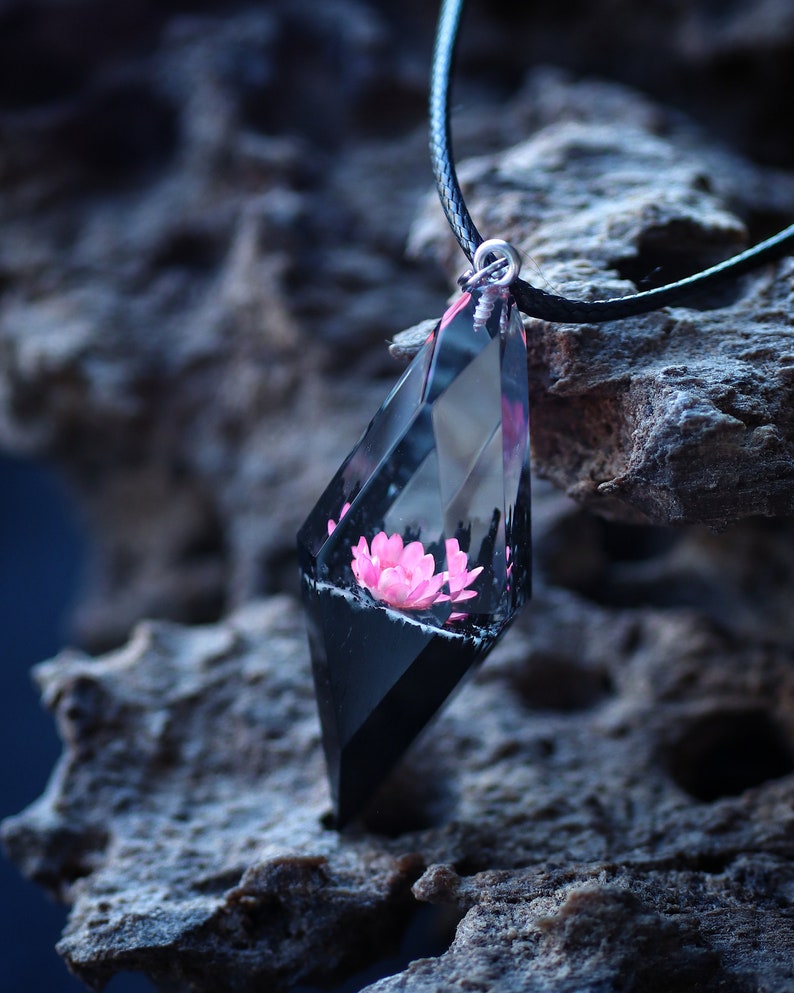 Op de afbeelding: Een geometrische hangerketting met een roze bloem ingekapseld in heldere hars met een zwarte basis. De hanger hangt aan een zwart koord. De ketting wordt getoond op een getextureerd, grijs oppervlak.