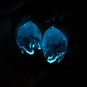 May include: Two glowing, faceted crystal pendants in a dark setting. The crystals emit a bright blue light, with internal structures visible. The pendants are suspended from small, dark loops, suggesting they are earrings or necklace charms.