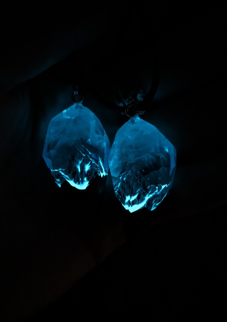 May include: Two glowing, faceted crystal pendants in a dark setting. The crystals emit a bright blue light, with internal structures visible. The pendants are suspended from small, dark loops, suggesting they are earrings or necklace charms.