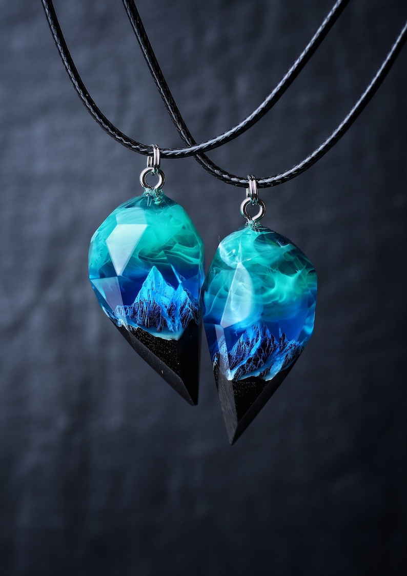 May include: Two matching pendant necklaces. Each pendant features a geometric shape with a black base, a mountain scene, and a translucent teal and blue resin top. The pendants hang from black cords.