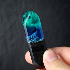 Può includere: Un ciondolo in resina blu scuro e verde con base in legno nero. La resina presenta un motivo a spirale e una silhouette di un lupo in piedi su una roccia.