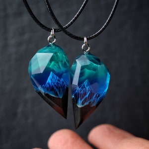 May include: Two matching resin pendants with a mountain scene, suspended on black cords. The pendants feature a faceted, clear top with blue and teal hues, transitioning to a dark base. The mountain design is visible within the resin.