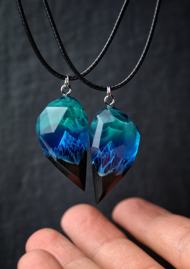 May include: Two matching resin pendants with a mountain scene, suspended on black cords. The pendants feature a faceted, clear top with blue and teal hues, transitioning to a dark base. The mountain design is visible within the resin.