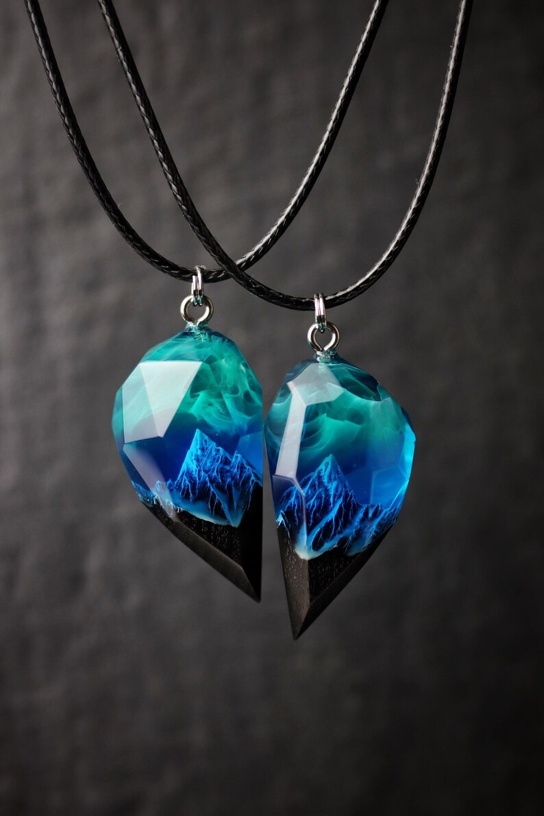 May include: Two matching pendant necklaces. Each pendant features a mountain scene encased in clear resin with blue and teal hues, set on a black wooden base. The pendants hang from black cords, creating a striking contrast against the dark background.