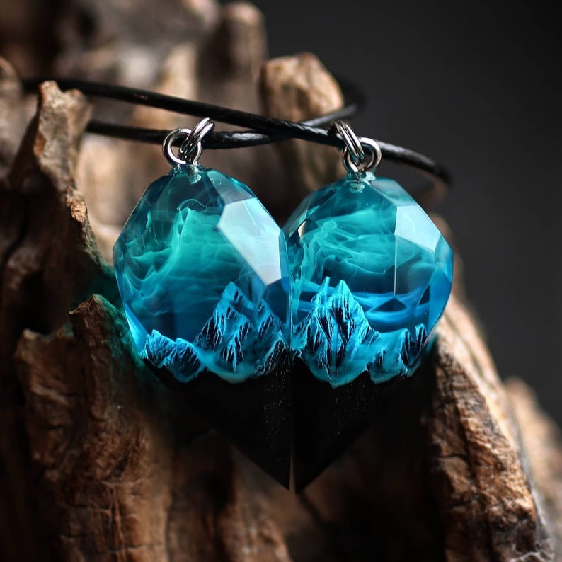May include: A pair of pendant necklaces featuring faceted, clear resin pendants with a blue and white swirling design, resembling clouds or water. The pendants are mounted on a black base with a mountain range design. The necklaces hang from a black cord.
