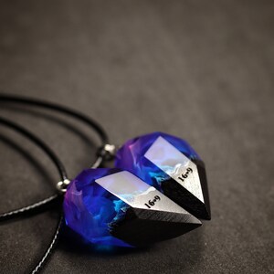 May include: Two matching pendant necklaces. Each pendant has a geometric shape with a black base and a translucent top filled with blue and purple hues. The text "16-9" is visible on the silver-coloured metal. The necklaces hang on black cords.