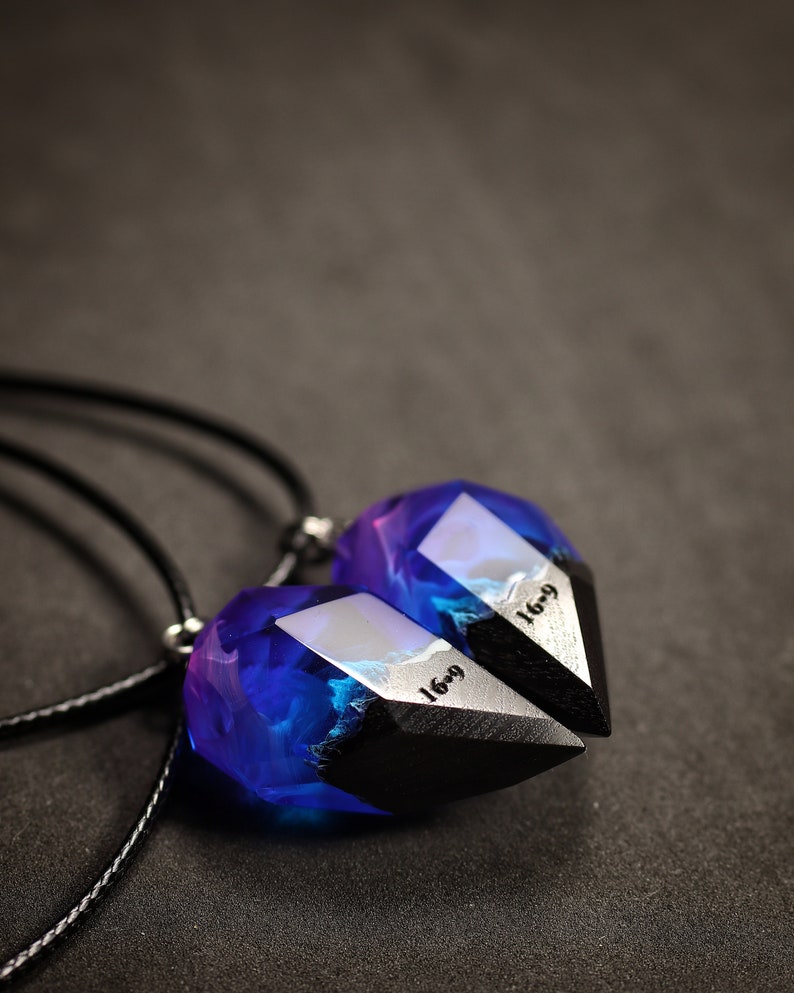 May include: Two matching pendant necklaces. Each pendant has a geometric shape with a black base and a translucent top filled with blue and purple hues. The text "16-9" is visible on the silver-coloured metal. The necklaces hang on black cords.