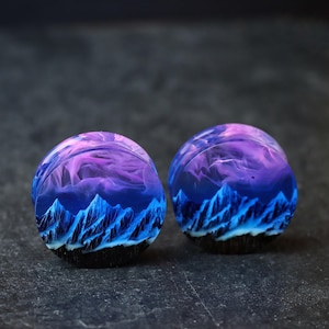 May include: A pair of round, wooden ear gauges with a blue and purple mountain scene inside. The gauges are made of resin and have a black outer edge.