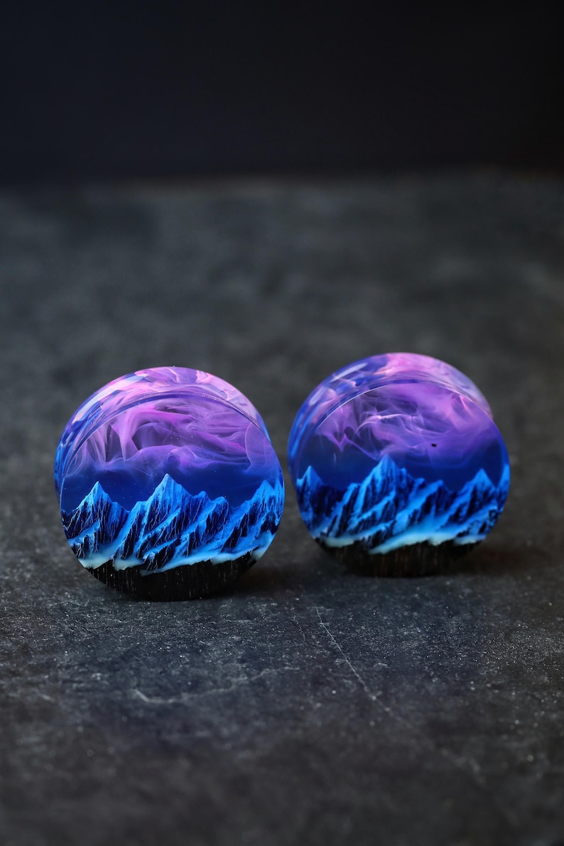 May include: A pair of round, wooden ear gauges with a blue and purple mountain scene inside. The gauges are made of resin and have a black outer edge.