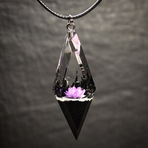 May include: A black cord necklace with a clear resin pendant shaped like a teardrop. The pendant contains a small purple flower and black and white details.