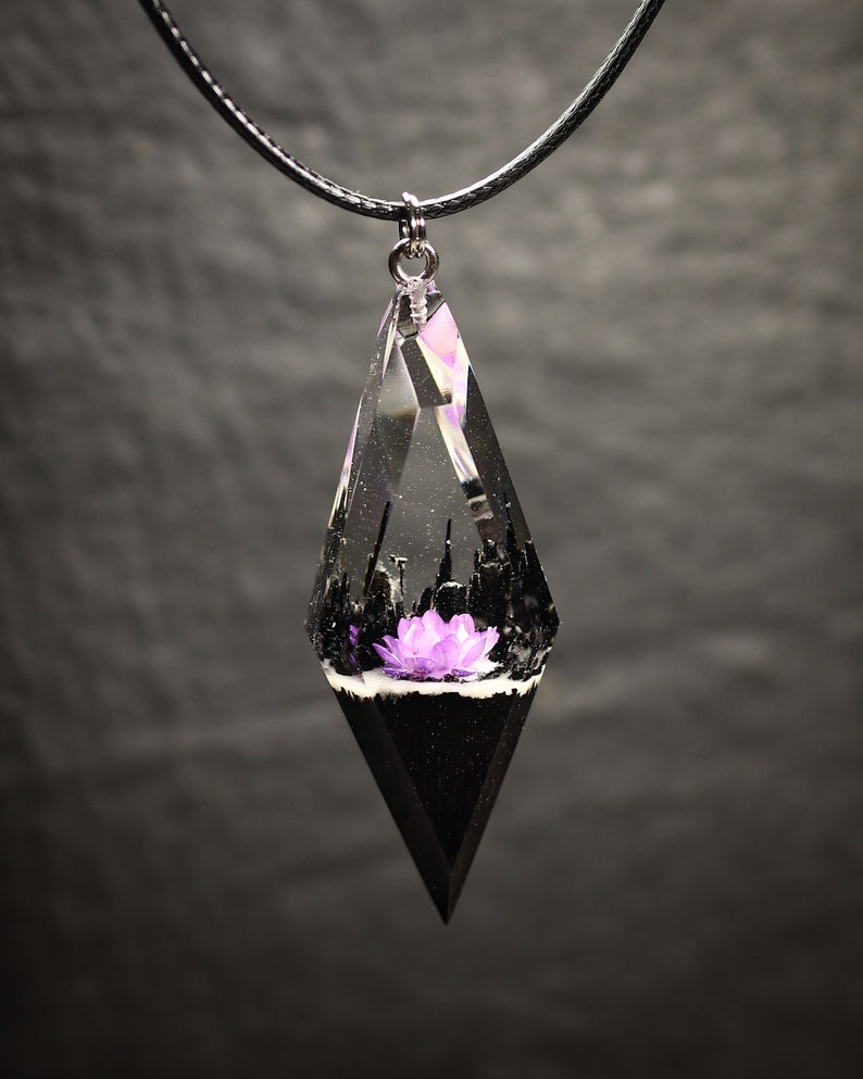 May include: A black cord necklace with a clear resin pendant shaped like a teardrop. The pendant contains a small purple flower and black and white details.