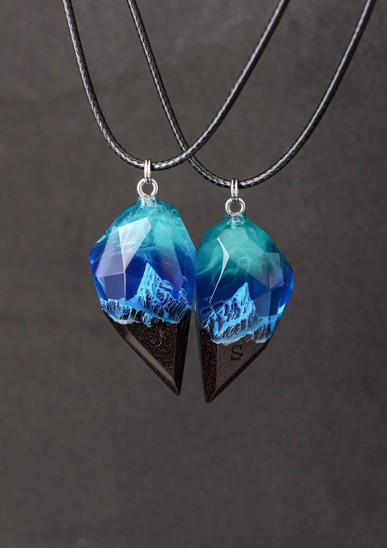 May include: Two matching pendant necklaces. Each pendant features a geometric shape with a dark brown base and a translucent blue and teal upper section, resembling a mountain range. The pendants hang from black cords.