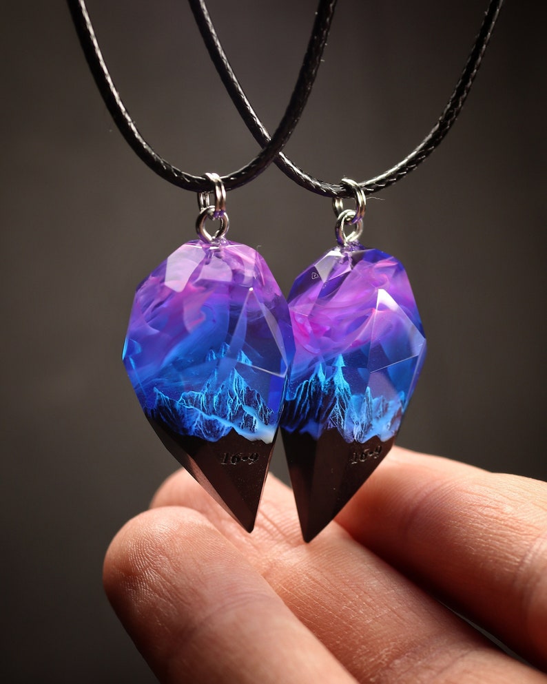 May include: Two matching pendant necklaces. Each pendant has a faceted, gemstone-like shape with a dark base and a translucent top filled with purple and blue hues. The pendants hang from black cords.