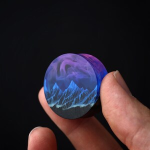 May include: A round, wooden plug with a blue and purple resin inlay featuring a mountain range and clouds. The plug is being held by a hand.