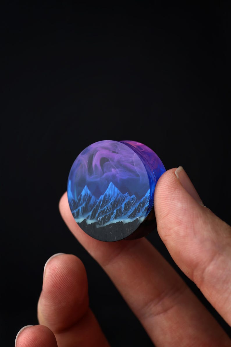 May include: A round, wooden plug with a blue and purple resin inlay featuring a mountain range and clouds. The plug is being held by a hand.