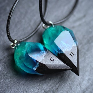 May include: Two matching heart-shaped pendants. Each pendant has a teal and blue crystal-like top and a black, pointed bottom. The pendants are connected to black cords. The letters "C" and "D" are visible on the black portion.