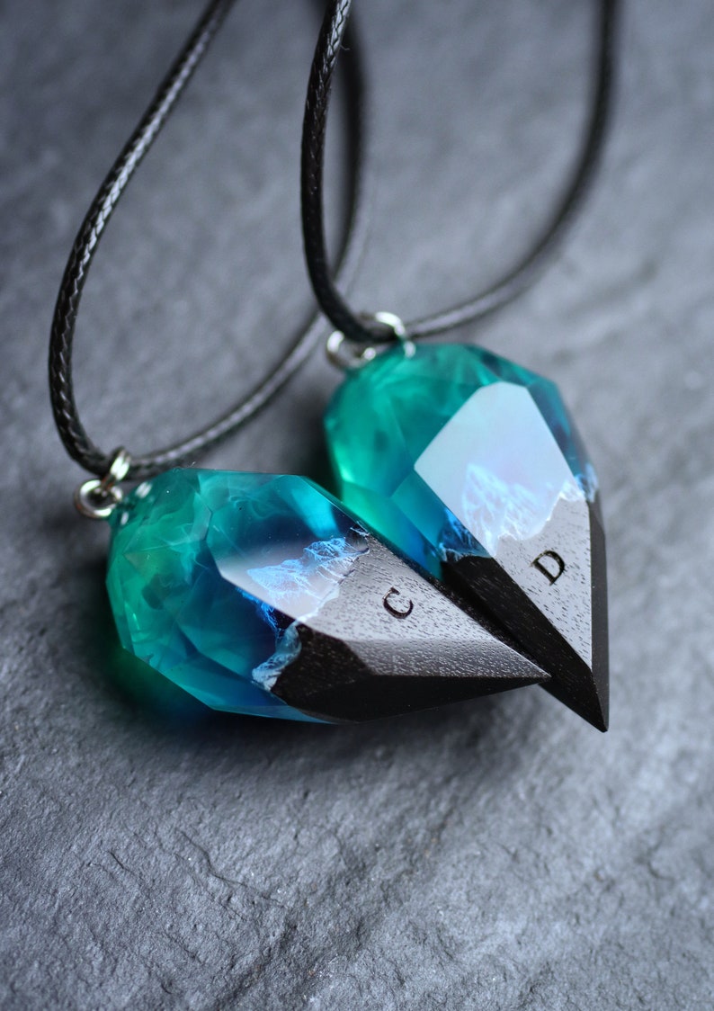 May include: Two matching heart-shaped pendants. Each pendant has a teal and blue crystal-like top and a black, pointed bottom. The pendants are connected to black cords. The letters "C" and "D" are visible on the black portion.