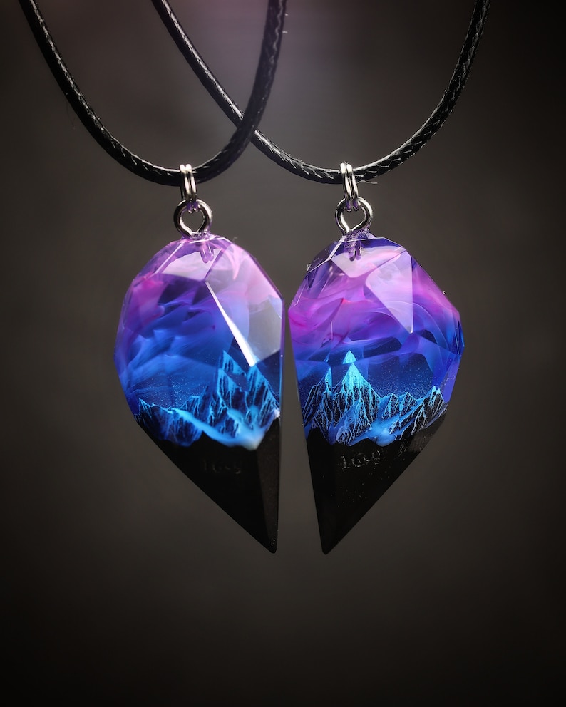 May include: Two matching pendant necklaces. Each pendant has a faceted, geometric shape with a black base and a translucent top. The top of each pendant has a gradient of purple and blue hues, with a mountain range design.