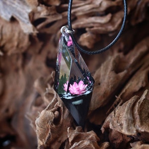 Op de afbeelding: Een unieke hangerketting met een heldere, facetgeslepen hars hanger met een roze bloem en een zwarte basis. De hanger hangt aan een zwart koord. De achtergrond is een getextureerd, bruin houten oppervlak.