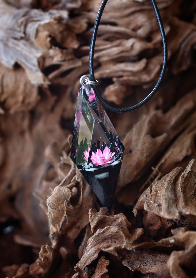 Op de afbeelding: Een unieke hangerketting met een heldere, facetgeslepen hars hanger met een roze bloem en een zwarte basis. De hanger hangt aan een zwart koord. De achtergrond is een getextureerd, bruin houten oppervlak.