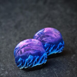 May include: A pair of purple and blue resin ear gauges with a mountain and sky design.