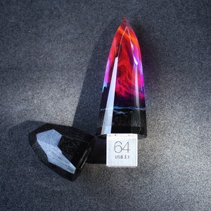 Puede incluir: Una unidad USB de resina negra y roja con forma de cristal puntiagudo. La unidad tiene una tapa de metal plateado con el texto "64 USB 3.1" impreso en ella.