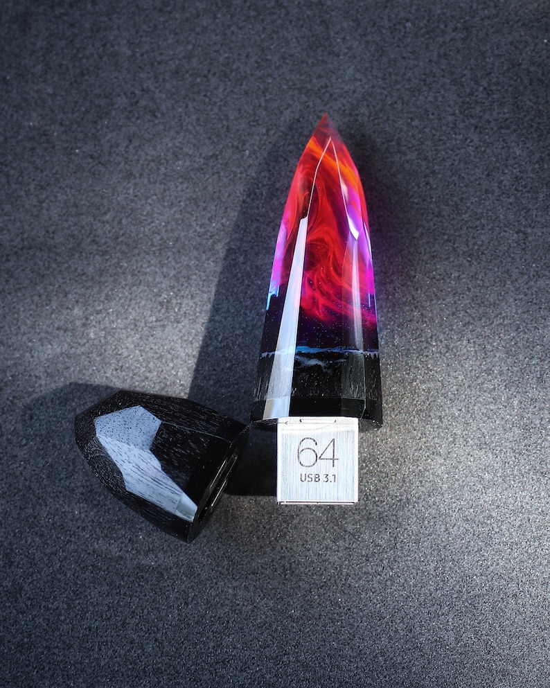 Puede incluir: Una unidad USB de resina negra y roja con forma de cristal puntiagudo. La unidad tiene una tapa de metal plateado con el texto "64 USB 3.1" impreso en ella.