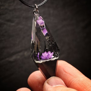 May include: A black and clear resin pendant necklace with a purple flower inside. The pendant is shaped like a teardrop and has a silver bail.