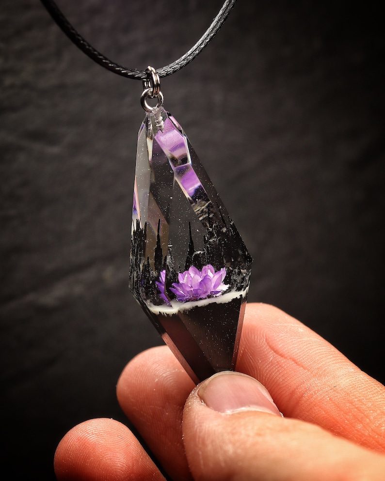 May include: A black and clear resin pendant necklace with a purple flower inside. The pendant is shaped like a teardrop and has a silver bail.