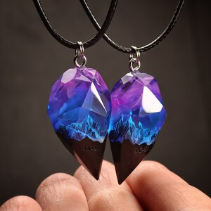 May include: Two matching pendant necklaces. The pendants are made of faceted resin, with a purple and blue gradient and a mountain scene. The bottom section is dark brown. The necklaces have black cords.