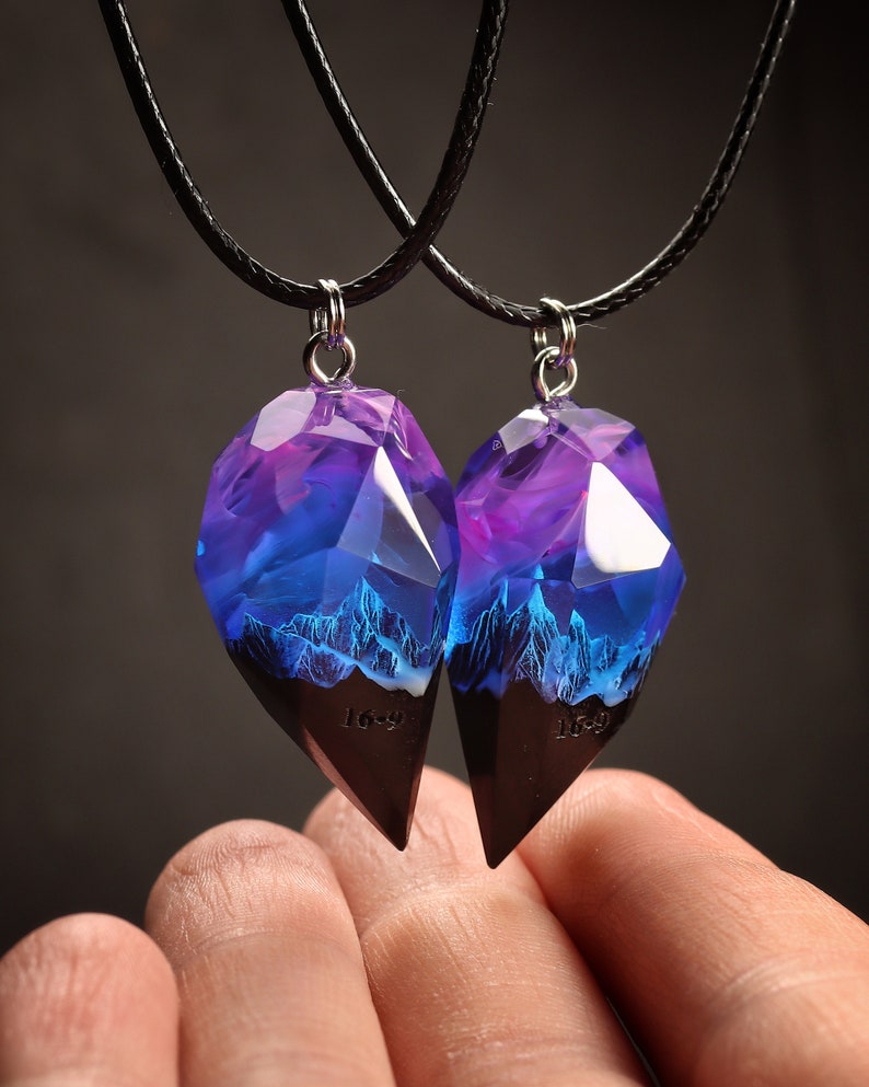May include: Two matching pendant necklaces. The pendants are made of faceted resin, with a purple and blue gradient and a mountain scene. The bottom section is dark brown. The necklaces have black cords.