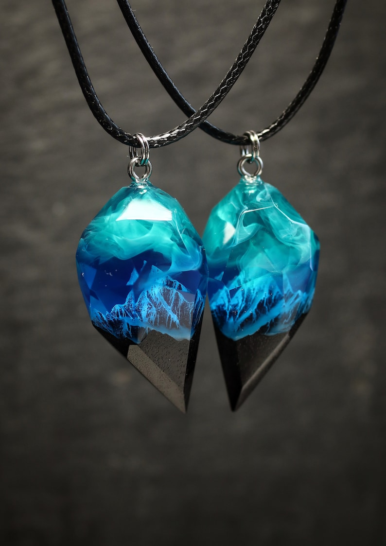 May include: Two matching pendant necklaces. Each pendant features a geometric shape with a black base and a clear resin top. The resin is filled with blue and white swirls and a mountain scene.