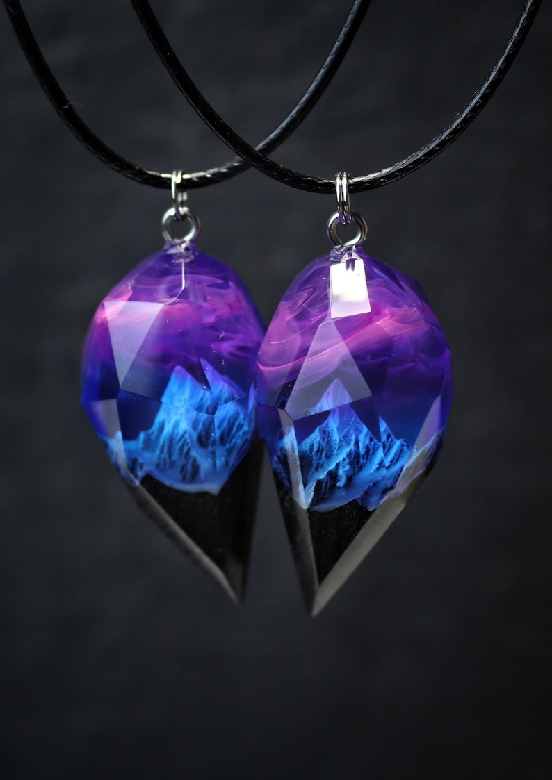 May include: Two matching pendant necklaces. Each pendant is a faceted, teardrop-shaped resin with a blue and purple landscape design. The bottom portion is black. The pendants hang from black cords with silver-coloured clasps.