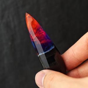 Puede incluir: Un bol&iacute;grafo puntiagudo de madera negra y oscura con una tapa de resina transparente llena de colores naranja, rojo y azul que se arremolinan.