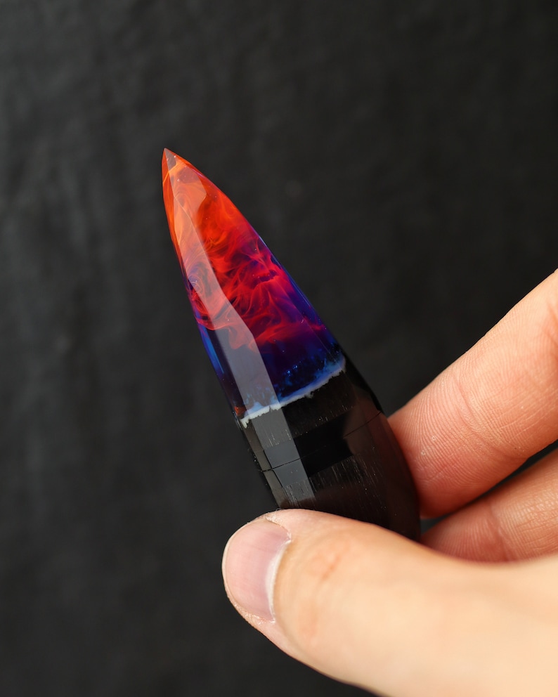 Puede incluir: Un bol&iacute;grafo puntiagudo de madera negra y oscura con una tapa de resina transparente llena de colores naranja, rojo y azul que se arremolinan.