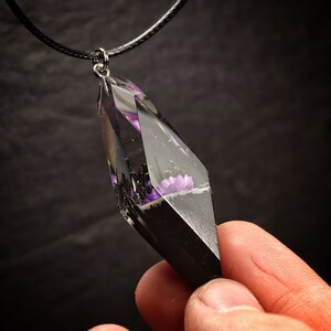 May include: A black cord necklace with a clear resin pendant featuring purple flowers encased in black resin. The pendant is shaped like a diamond.
