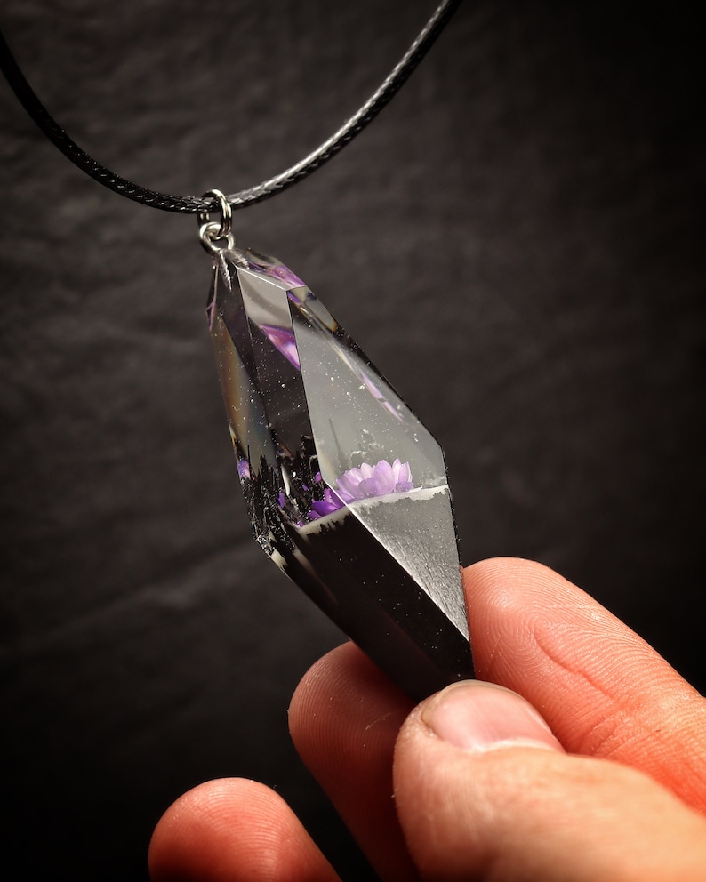 May include: A black cord necklace with a clear resin pendant featuring purple flowers encased in black resin. The pendant is shaped like a diamond.