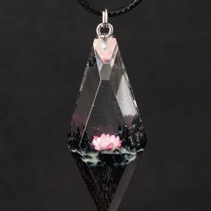 Op de afbeelding: Een heldere, ruitvormige hanger met een zwart koord. De hanger bevat een miniatuurlandschap met een roze lotusbloem, donkere bomen en een witte, mistige basis. De hanger hangt tegen een zwarte achtergrond.