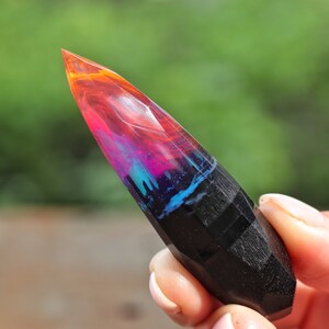 Puede incluir: Una punta de cristal de resina negra y colorida con un patr&oacute;n de remolino de rosa, naranja, azul y negro. El cristal es puntiagudo en la parte superior y tiene una superficie lisa y pulida.
