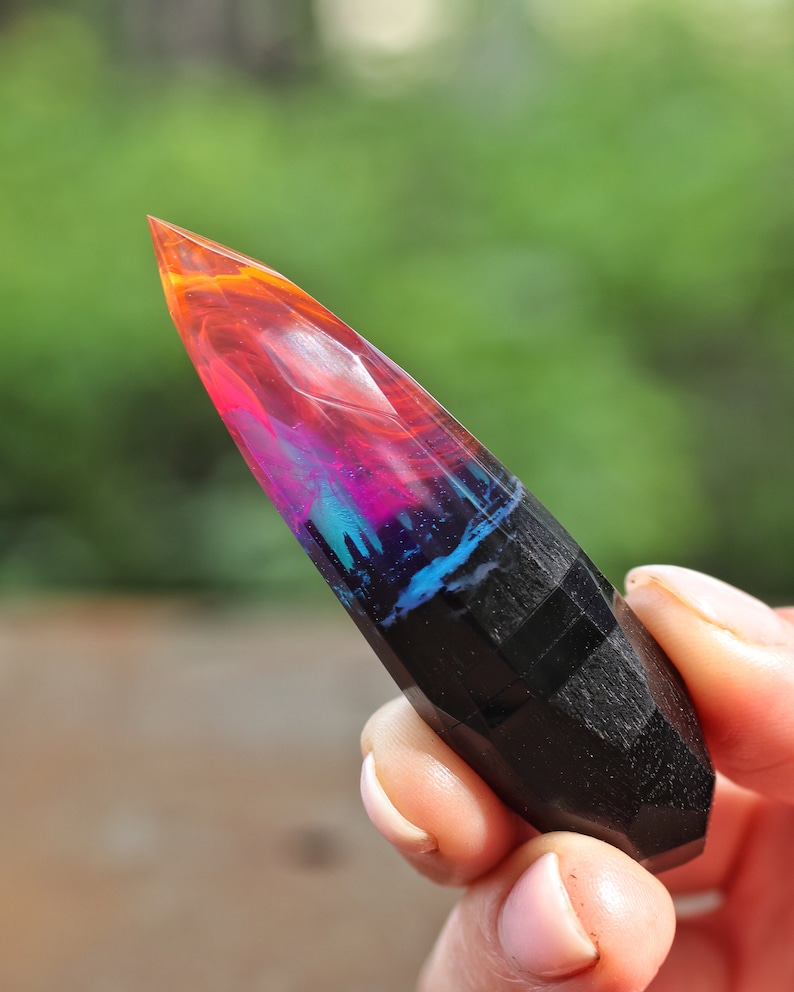 Puede incluir: Una punta de cristal de resina negra y colorida con un patr&oacute;n de remolino de rosa, naranja, azul y negro. El cristal es puntiagudo en la parte superior y tiene una superficie lisa y pulida.