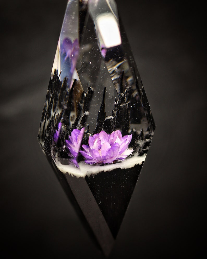 May include: A clear, diamond-shaped pendant with a purple flower encased in resin. The flower is surrounded by black resin and white accents.