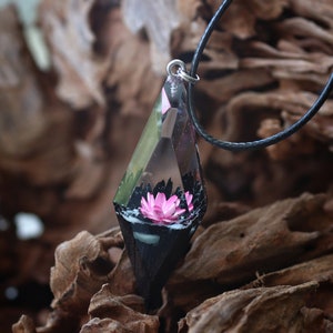 Op de afbeelding: Een ketting met een hanger van heldere, geometrische hars. In de hanger zit een roze lotusbloem. De hanger hangt aan een zwart koord. De achtergrond is wazig hout.