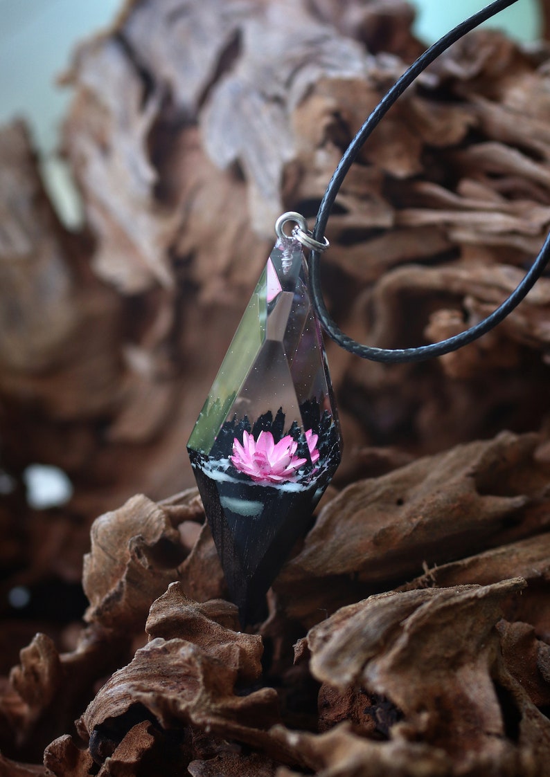 Op de afbeelding: Een ketting met een hanger van heldere, geometrische hars. In de hanger zit een roze lotusbloem. De hanger hangt aan een zwart koord. De achtergrond is wazig hout.