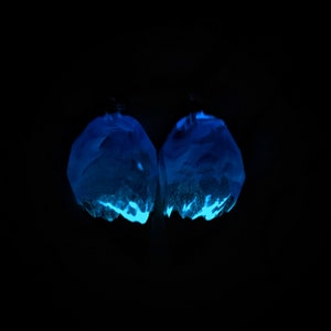 May include: Two glowing blue earrings against a black backdrop. The earrings are irregularly shaped with facets, and emit a bright, neon-like blue glow. The bottom edges have a lighter, textured appearance, creating a contrast.