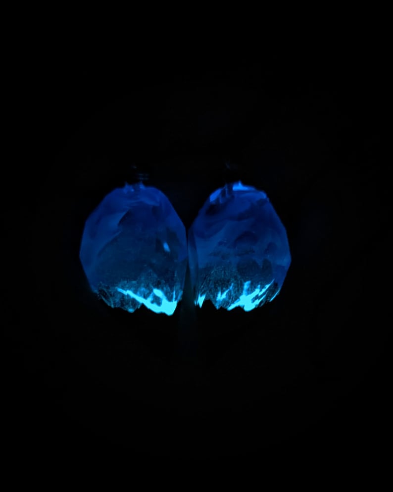 May include: Two glowing blue earrings against a black backdrop. The earrings are irregularly shaped with facets, and emit a bright, neon-like blue glow. The bottom edges have a lighter, textured appearance, creating a contrast.