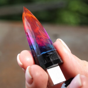 Puede incluir: Una unidad USB de resina colorida y multicapa con forma de punta de cristal. La unidad es negra en la base y tiene un conector USB plateado.